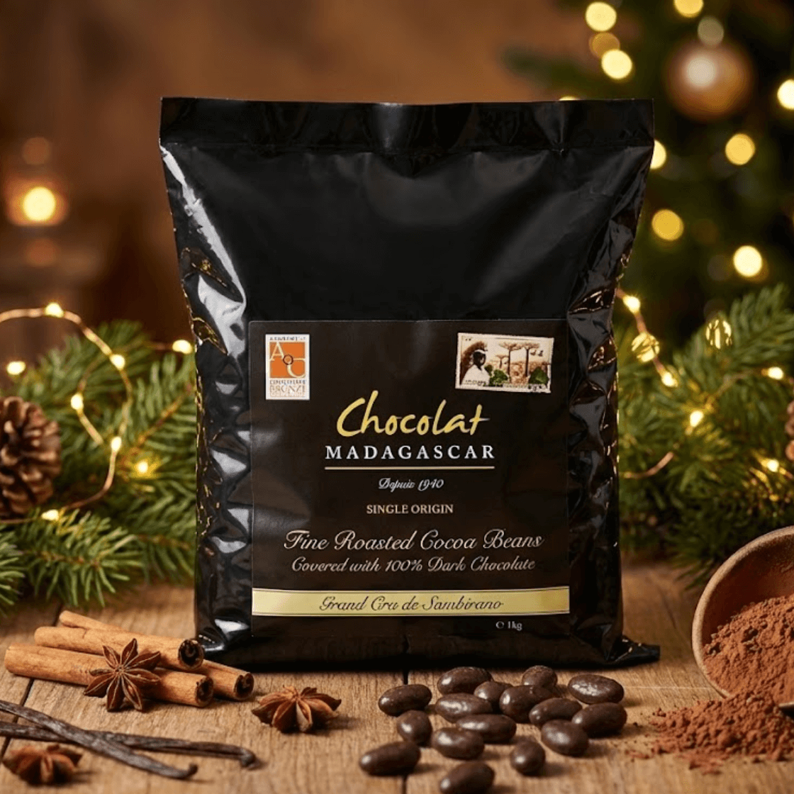 Chocolat Madagascar single-origin fine roasted cocoa beans covered in 100% dark chocolate, presented in a black 1 kg package on a wooden table with cocoa beans, cocoa powder, cinnamon sticks, star anise, and festive greenery in a warm, cozy setting.